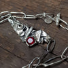 silver necklace with rough texture and oxidized pendant with red garnet. All the links are oval different shaped and handmade. theres also two long rough silver rectangular beads. Closes with a organic silver toggle, has one little organic silver charm at the end