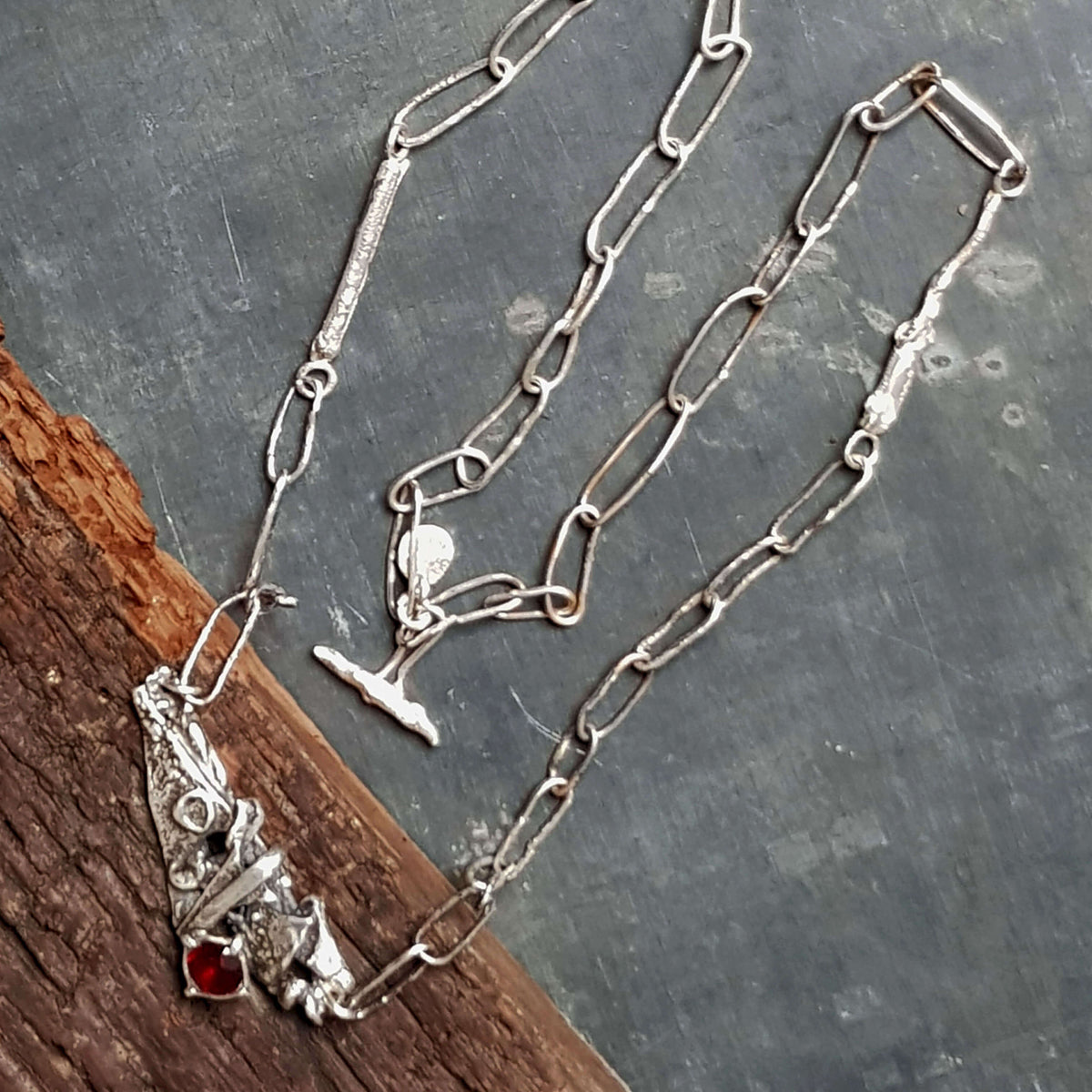 rustic looking long handmade necklace, hand made silver links, closes with a handmade silver toggle, featuring a rough textured silver pendant in a brutalist style with a red garnet gemstone